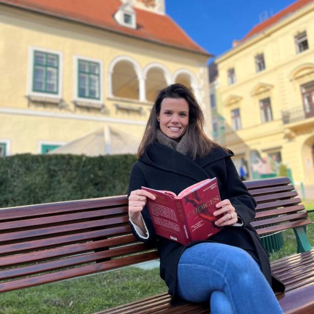 Autorin Katharina Riha sitzt mit ihrem aufgeschlagenen Buch in den Händen auf einer Parkbank.