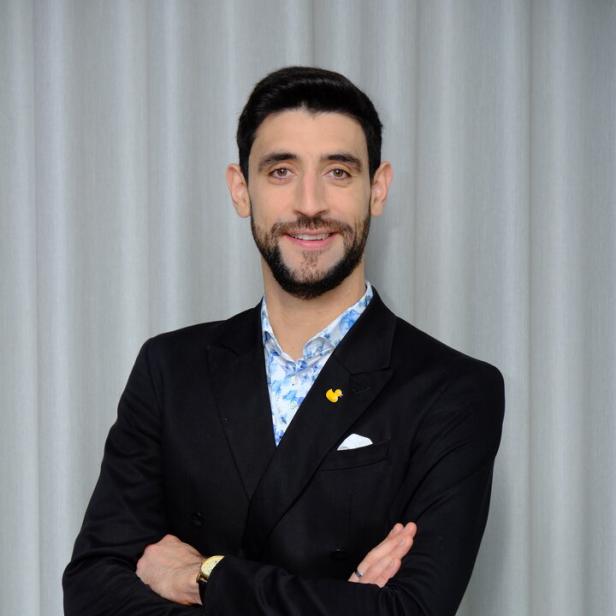 Ein Mann mit dunklem Haar und Bart trägt einen schwarzen Anzug und steht mit verschränkten Armen vor einem hellen Vorhang.