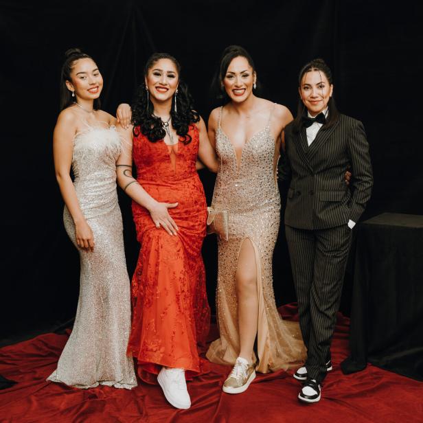 Vier elegant gekleidete Frauen posieren gemeinsam, drei in Abendkleidern und eine im Anzug, vor schwarzem Hintergrund.