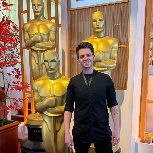 Bernhard Zimmerl steht vor drei menschengroßen Oscar-Figuren und lächelt im Koch-Outfit in die Kamera.