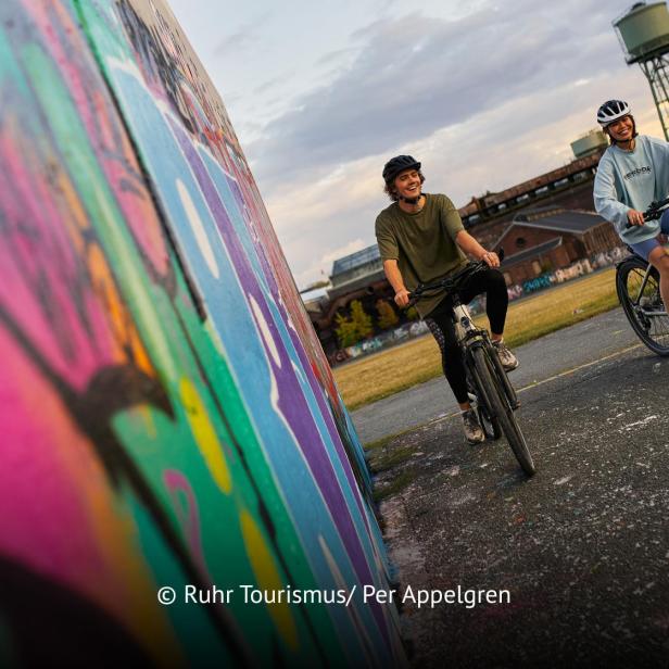 Zwei Radfahrer fahren an einer bunt besprühten Wand im urbanen Ruhrgebiet entlang.