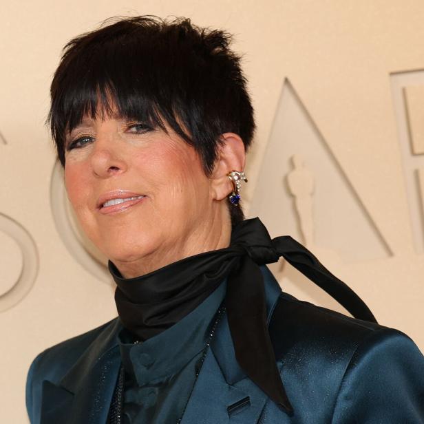 Diane Warren mit kurzem dunklem Haar, Ohrschmuck und schwarzer Schleife um den Hals vor hellem Hintergrund.