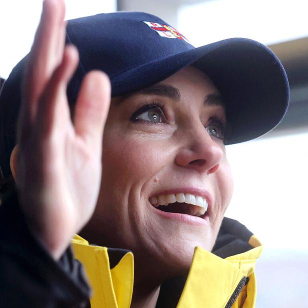 Prinzessin Kate lächelt, trägt eine blaue Kappe und eine gelbe Jacke und winkt.