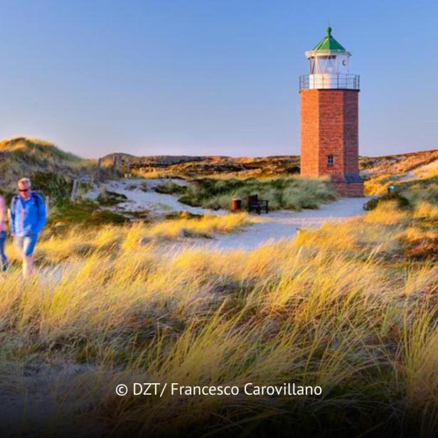 Zwei Personen spazieren bei Sonnenuntergang durch Dünenlandschaft mit kleinem Leuchtturm auf Sylt.