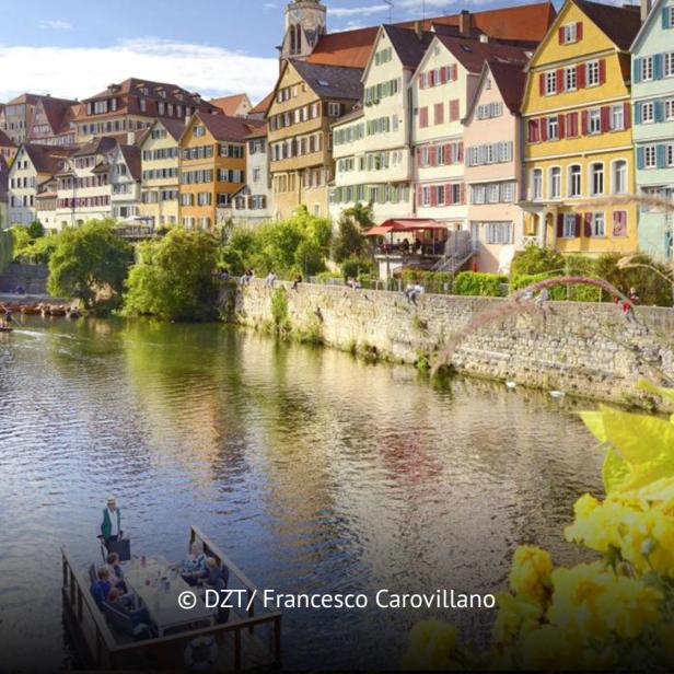 Bunte Fachwerkhäuser am Flussufer mit Stocherkahnfahrten und blühenden Blumen im Vordergrund.