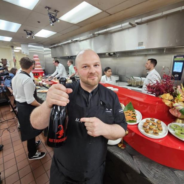 Ein Koch hält eine große schwarze Flasche mit rotem Skorpion-Logo in einer Restaurantküche, im Hintergrund arbeiten weitere Köche.