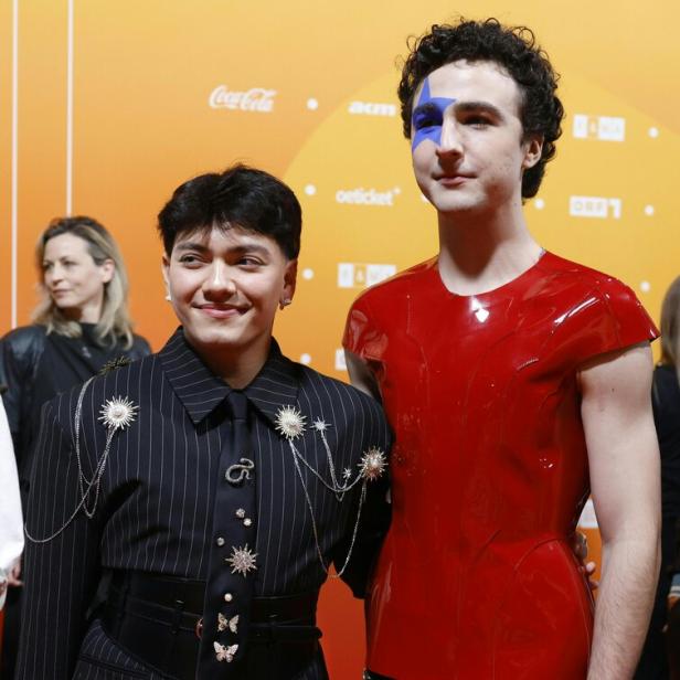 Zwei Personen posieren vor orangefarbenem Hintergrund, eine trägt einen Nadelstreifenanzug mit Schmuck, die andere ein rotes Oberteil und Gesichtsbemalung.