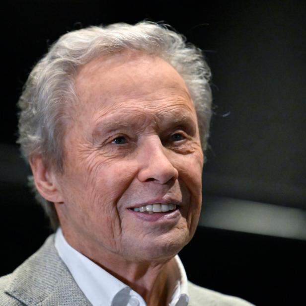 Ein Mann mit grauem Haar lächelt und trägt ein helles Jackett vor dunklem Hintergrund.