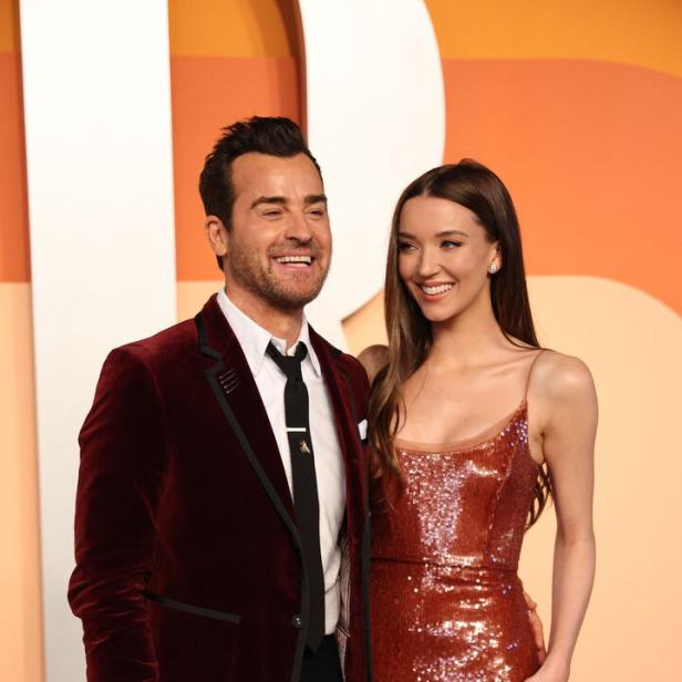 Justin Theroux im Samtjackett und Nicole Brydon Bloom im Paillettenkleid lächeln vor einem farbigen Hintergrund.