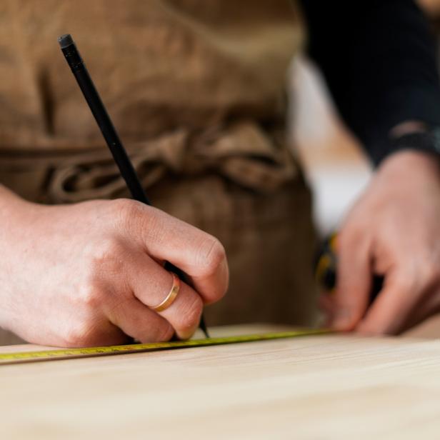 Hände messen und markieren ein Holzbrett mit Bleistift und Maßband in einer Werkstatt.