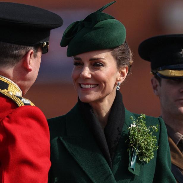 Eine elegant gekleidete Prinzessin Kate in Grün mit Hut und Anstecksträußchen spricht lächelnd mit zwei uniformierten Männern.