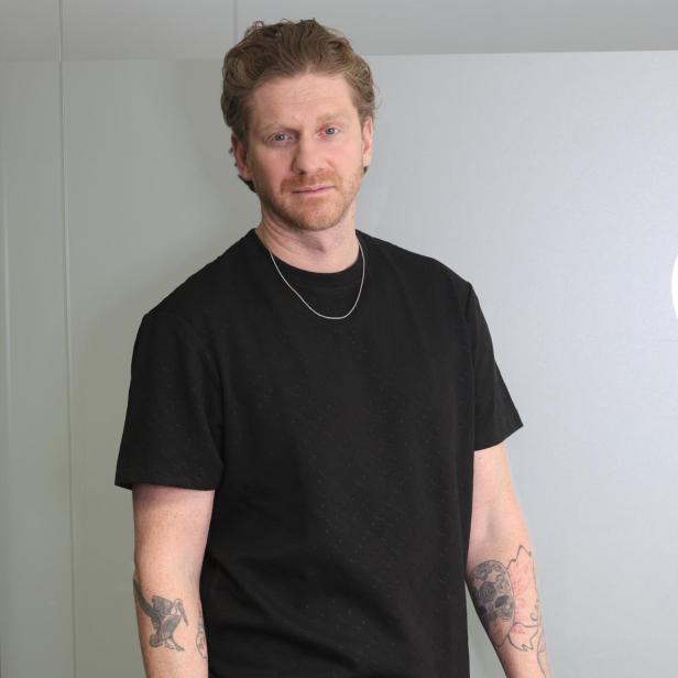 Ein Mann mit lockigen Haaren trägt ein schwarzes T-Shirt, helle Hose und zeigt mehrere Tattoos an den Armen.