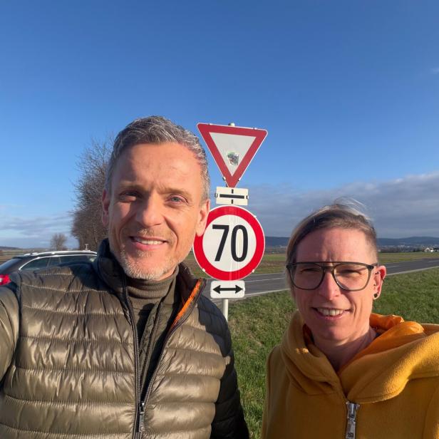 Zwei Personen stehen bei Sonnenschein neben einer Landstraße vor einem Vorfahrt-achten- und einem 70-km/h-Schild.