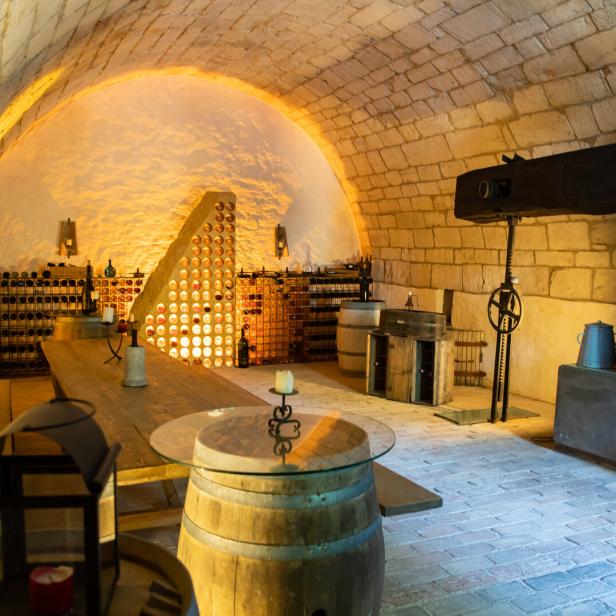 Gewölbekeller mit Weinregalen, Holztischen, Fässern und stimmungsvoller Beleuchtung.