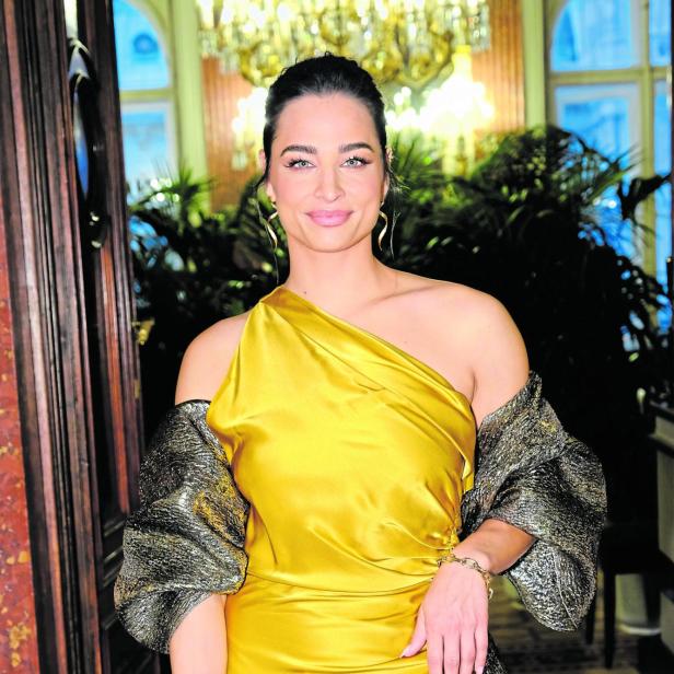 Amira Aly in goldenem, schulterfreiem Kleid mit glitzerndem Schal und Clutch.