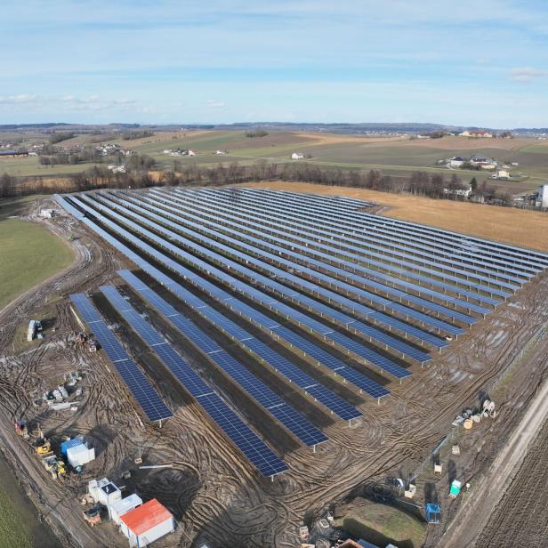 Große Solaranlage mit zahlreichen Solarpanels auf einem Feld, umgeben von ländlicher Landschaft und vereinzelten Gebäuden.