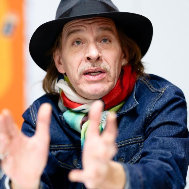 Mann mit Hut, bunter Schal und Jeansjacke gestikuliert mit den Händen und spricht, im Hintergrund unscharfe Farben.