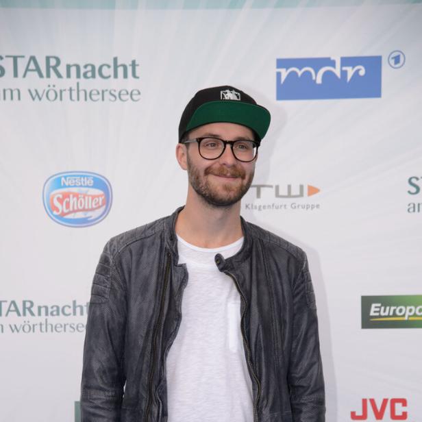 Mark Forster mit Brille, Bart, schwarzer Kappe, grauer Lederjacke und weißem T-Shirt steht vor einer Sponsorenwand.