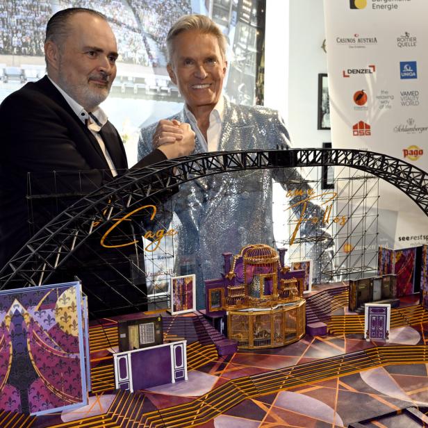 Zwei Männer posieren gemeinsam vor einem detailreichen, bunten Bühnenmodell mit goldenen und violetten Elementen.