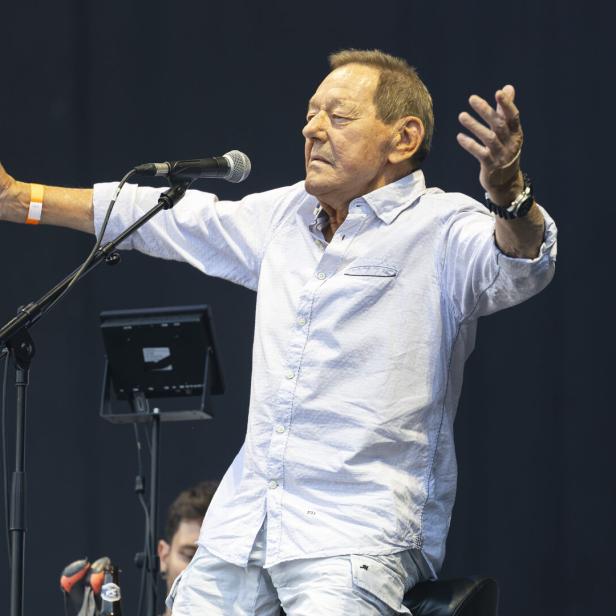 Ein Mann mit ausgebreiteten Armen singt an einem Mikrofon auf einer Bühne vor dunklem Hintergrund.