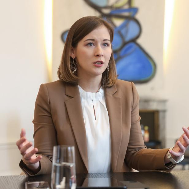 Eine Frau mit braunen Haaren und beigem Blazer spricht gestikulierend an einem Tisch in einem modernen Büro.