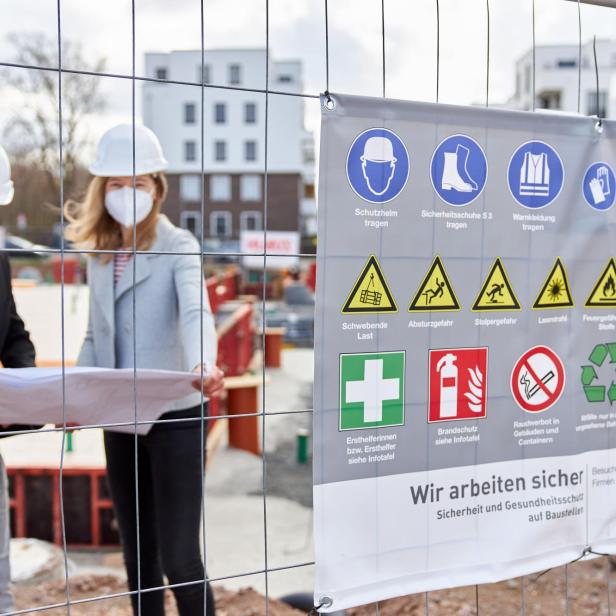 Zwei Personen mit Schutzhelmen und Masken betrachten Baupläne hinter einem Bauzaun mit Warnhinweisen.