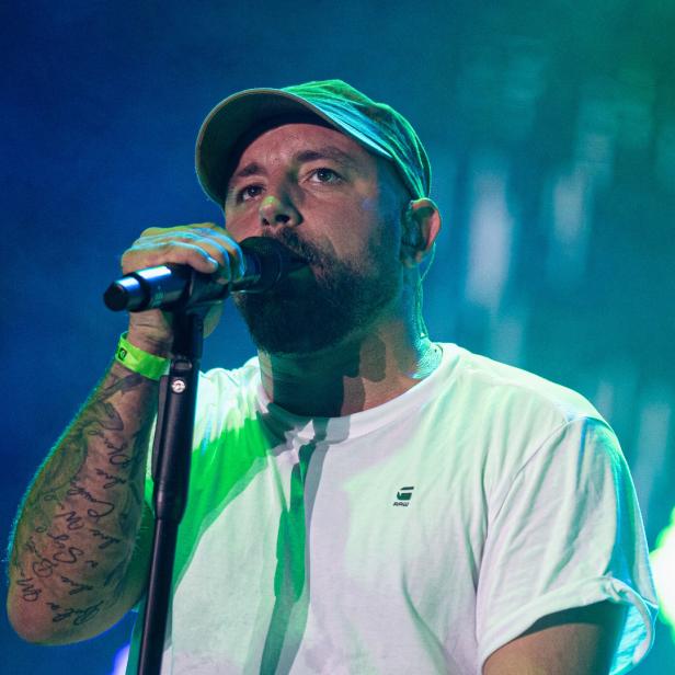 Ein bärtiger Christopher Seiler mit Kappe und Tattoos singt in ein Mikrofon vor blau-grünem Hintergrund.
