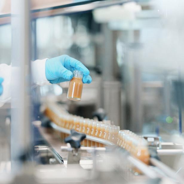 Eine Person mit blauen Handschuhen hält ein kleines Glasfläschchen an einem Fließband.