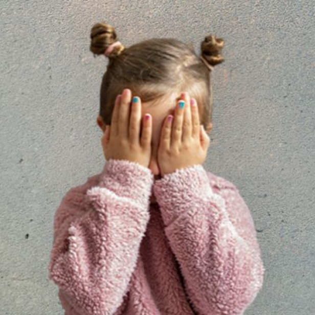 Kind verdeckt das Gesicht mit den Händen vor einer grauen Wand.