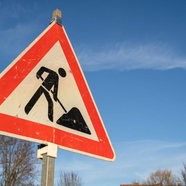 Verkehrsschild mit rotem Rand warnt vor einer Baustelle, im Hintergrund blauer Himmel und Bäume.