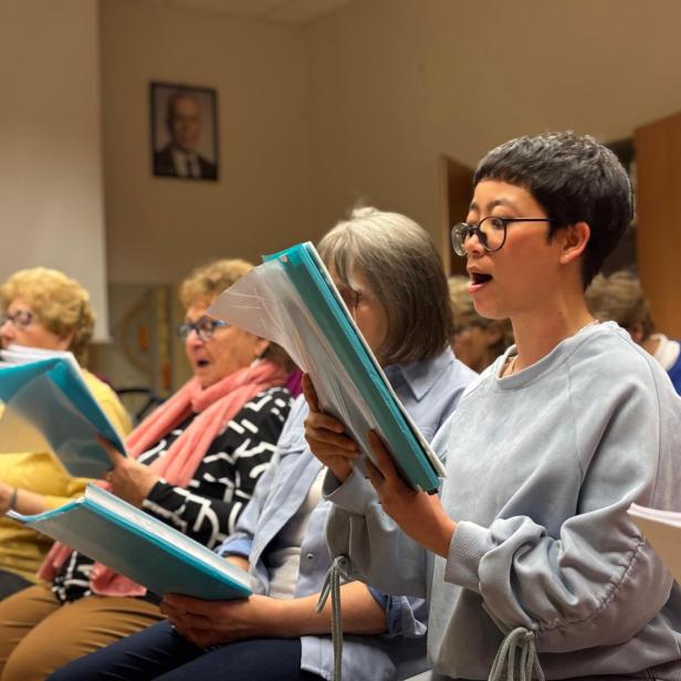 Eine Aufnahme von einer Chorprobe, mehrere ältere Personen sind mit Mappen beim Singen zu sehen. Im Fokus steht eine junge Frau aus Vietnam.