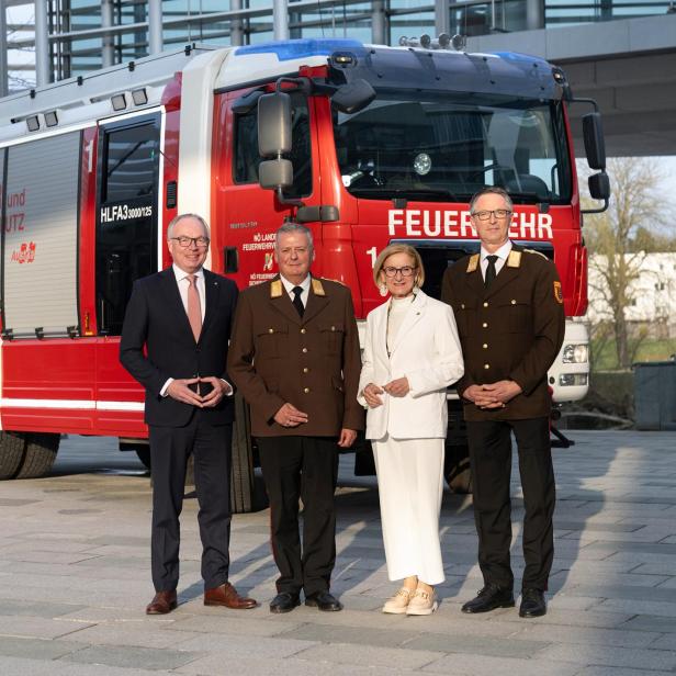 Vier Personen stehen vor einem roten Feuerwehrfahrzeug, drei in Uniform und eine Person im weißen Anzug.
