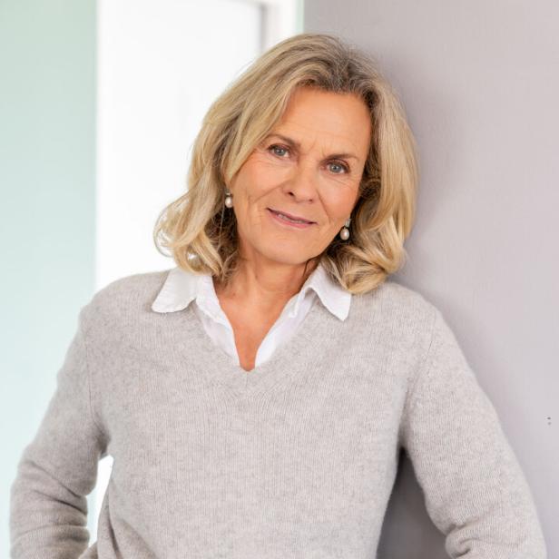 Eine Frau mit blonden Haaren, trägt einen grauen Pullover und Jeans, lehnt an einer Wand.