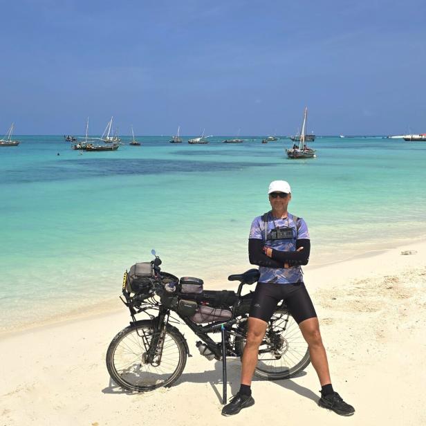 Der Weltumradler posiert mit seinem Fahrrad am Strand, im Hintergrund türkisfarbenes Wasser und Boote.