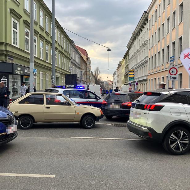 Mehrere Autos und Polizisten stehen an einer Straßensperre in einer Stadt mit Wohnhäusern und einem FPÖ-Stand.
