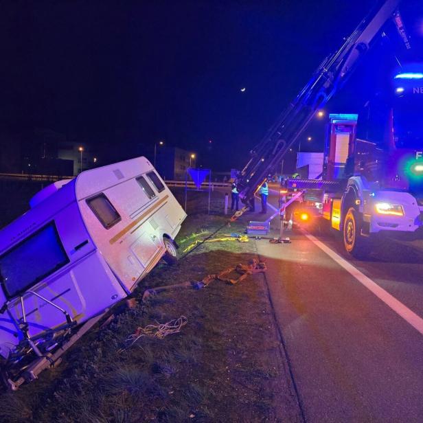 Ein Wohnwagen liegt schräg im Straßengraben, während ein Feuerwehrfahrzeug mit Kran ihn bei Nacht sichert.