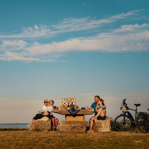 Vier Personen sitzen an einem steinernen Tisch im Freien, trinken Wein und genießen ein Picknick, Fahrräder stehen daneben.