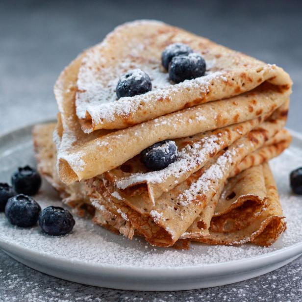Mehrere zusammengefaltete Crêpes mit Puderzucker bestreut und mit frischen Heidelbeeren garniert auf einem Teller.
