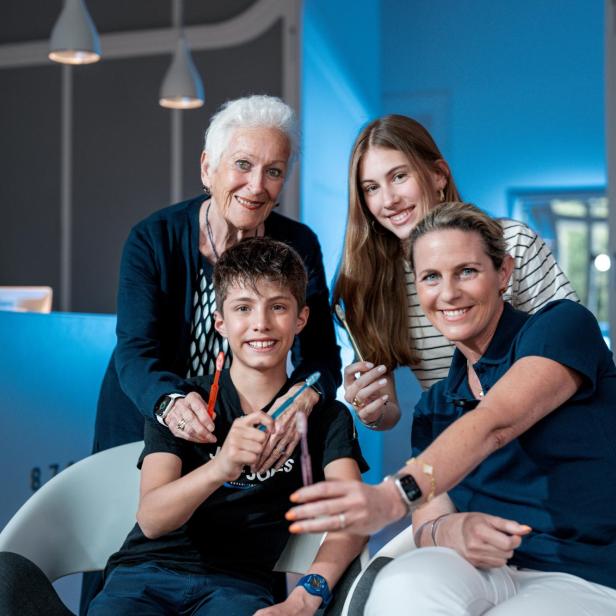 Vier Personen unterschiedlichen Alters halten gemeinsam Zahnbürsten und lächeln in die Kamera.