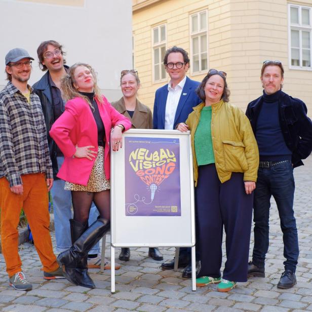 Mehrere bunt gekleidete Menschen, darunter Neubau Bezirksvorsteher Reiter, stehen vor einem Plakatschild auf Pflastersteinen