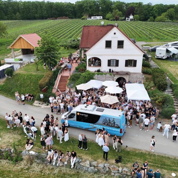 Viele Menschen feiern vor einem weißen Haus mit roten Dachziegeln, umgeben von Weinbergen und mehreren weißen Pavillons.
