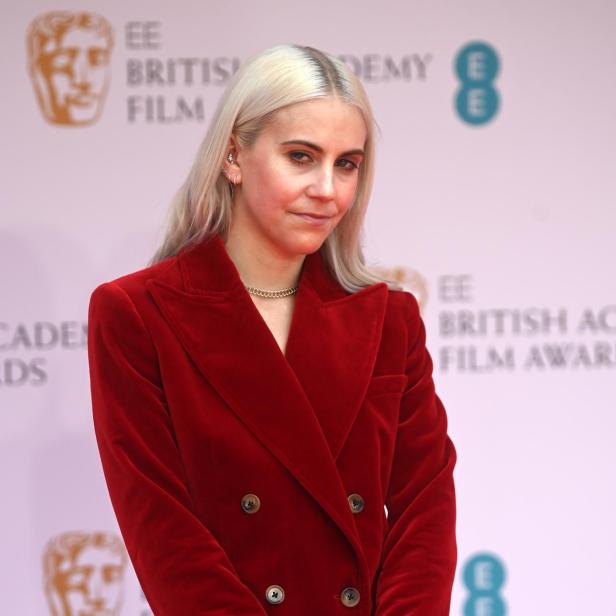 India Rose James mit blonden Haaren trägt einen roten Samtanzug vor dem Hintergrund der BAFTA Film Awards.