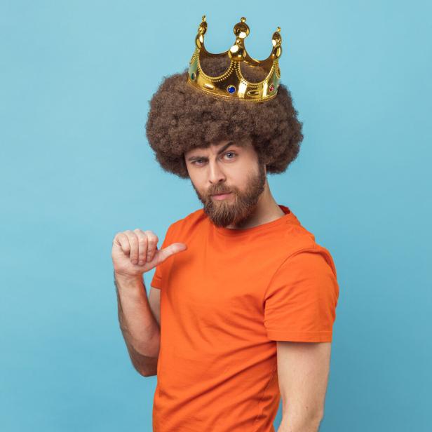 Mann mit Lockenperücke und goldener Krone trägt ein orangefarbenes T-Shirt und zeigt mit dem Daumen auf sich selbst.