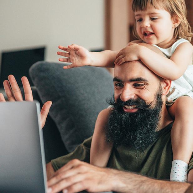 Ein Mann lernt mit einem Laptop zu Hause, während ein kleines Kind auf seinen Schultern sitzt und beide lächeln.