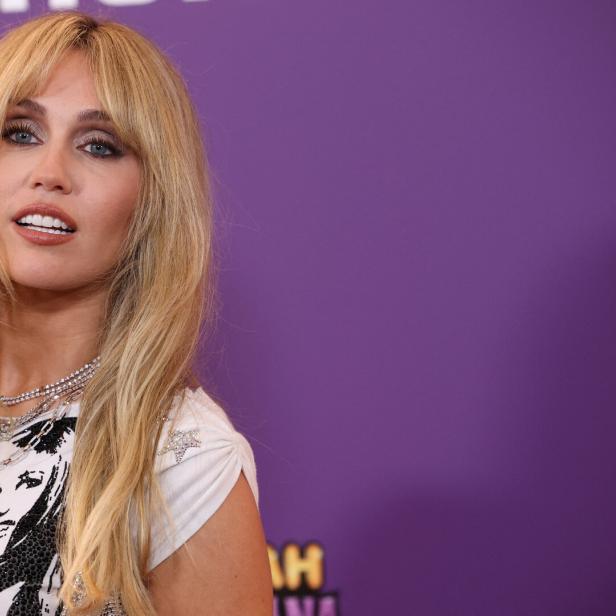 Miley Cyrus mit blonden Haaren und Halsketten vor lila Hintergrund.