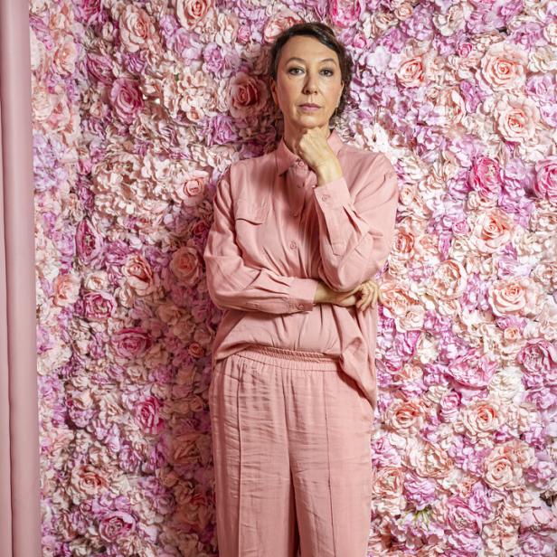 Frau in rosa Outfit steht vor einer Wand aus rosa und pinken Kunstblumen und schaut nachdenklich.