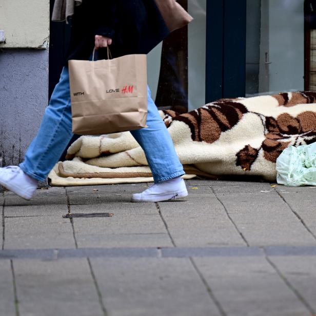 Eine Person liegt unter einer Decke auf dem Gehweg, daneben Einkaufstüten, während jemand vorbeigeht.