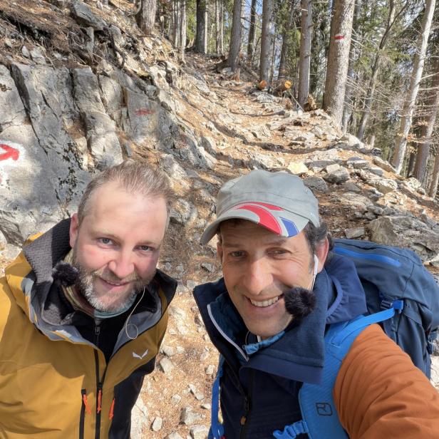 Josh. und Tom Walek wandern gemeinsam auf einem steinigen Waldweg mit markiertem Wanderzeichen.