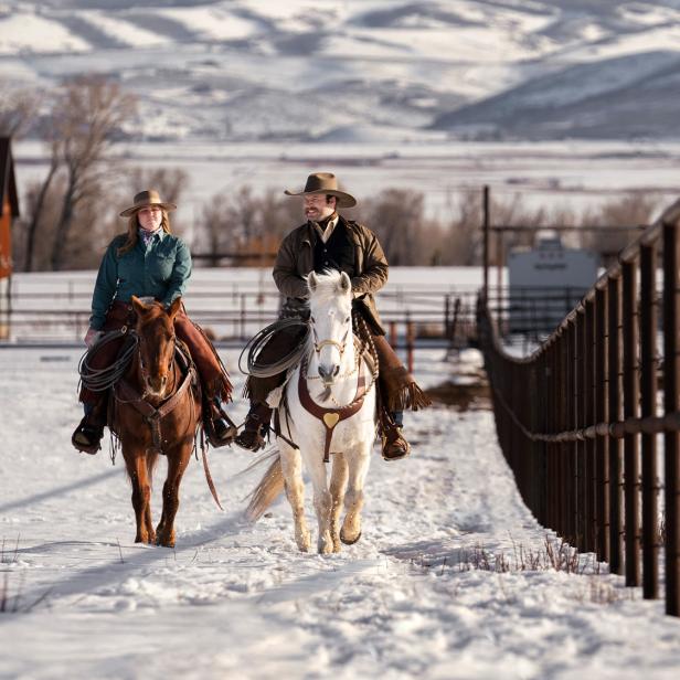 Zwei Reiter reiten auf verschneitem Reitplatz neben einem Zaun, im Hintergrund Berge und Stallgebäude.