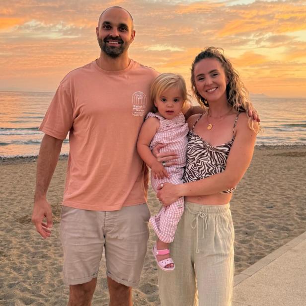 Gus und Emily Forrester stehen mit ihrer Tochter Leni am Strand und blicken gemeinsam in die Kamera, während die Sonne untergeht.